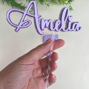 Könnte beinhalten: Ein lila Tortenaufsatz mit dem Namen "Amelia" in Schreibschrift. Die Buchstaben sind mit silbernem Glitzer umrandet, und der Aufsatz wird von einem durchsichtigen Kunststoffstab gehalten.