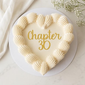 May include: A heart-shaped cake with smooth off-white icing, decorated with piped cream borders. The centre features the text "Chapter 30" in a glittery gold script font. The cake is presented on a round white base.