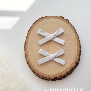 May include: Two white fabric hair bows on a wooden slice. The bows are tied in a simple knot and are arranged in an X shape.