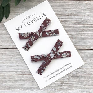 May include: Two brown floral hair bows with white flowers. The bows are on a white card with the text "MY LOVELLIE" and website information.