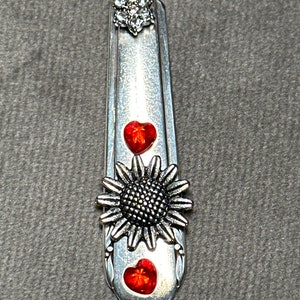 May include: A silver spoon necklace with a red heart, a sunflower, and a crystal flower charm.