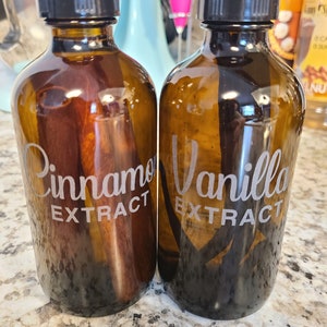 May include: Two glass bottles with black lids and labels. The bottle on the left is filled with a dark liquid and has a cinnamon stick inside. The label reads "Cinnamon Extract". The bottle on the right is filled with a dark liquid and has a vanilla bean inside. The label reads "Vanilla Extract".
