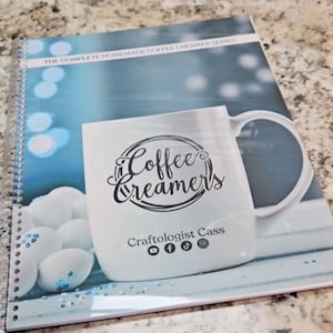May include: A blue and white spiral bound notebook with a white coffee mug on the cover. The mug has the text "Coffee Creamers" and "Craftologist Cass" with social media icons. The background is a blue and white blurred image of a winter scene.