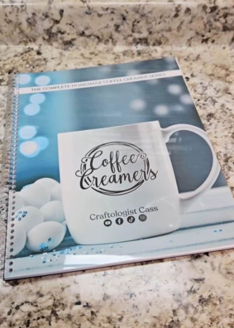 May include: A blue and white spiral bound notebook with a white coffee mug on the cover. The mug has the text "Coffee Creamers" and "Craftologist Cass" with social media icons. The background is a blue and white blurred image of a winter scene.