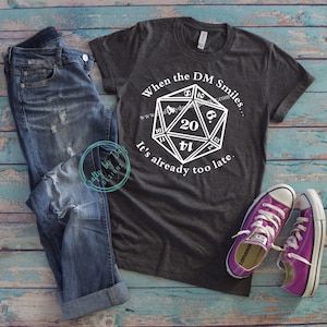 May include: A gray t-shirt with a white graphic of a twenty-sided die and the text "When the DM Smiles... It's already too late."