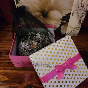 May include: A pink gift box with a white lid featuring gold polka dots and a pink ribbon. The box is open and contains a black mesh bag filled with a jigsaw puzzle.