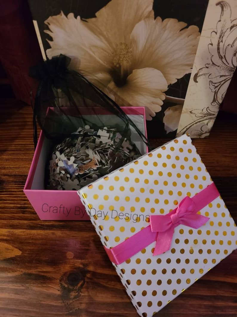 May include: A pink gift box with a white lid featuring gold polka dots and a pink ribbon. The box is open and contains a black mesh bag filled with a jigsaw puzzle.