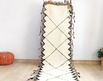 May include: A white Moroccan rug with black diamond patterns and fringe. The rug is hanging on a white wall.