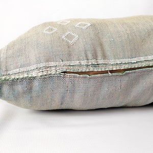 May include: A rectangular decorative pillow in shades of gray, blue, and beige. The pillow features white diamond-shaped embroidery and a brown zipper. The edges are trimmed with green stitching and a white tassel.