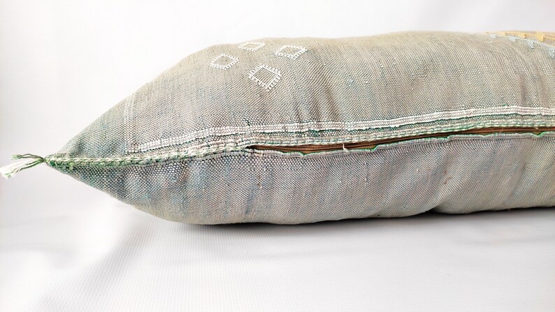 May include: A rectangular decorative pillow in shades of gray, blue, and beige. The pillow features white diamond-shaped embroidery and a brown zipper. The edges are trimmed with green stitching and a white tassel.