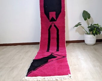 Puede incluir: Una alfombra larga y estrecha de color rosa y negro con un patrón geométrico. La alfombra tiene un borde de flecos y está hecha de lana.