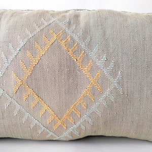 May include: Rectangular decorative pillow in a neutral gray color. The pillow features a central diamond pattern in gold and light blue, with additional geometric designs. The pillow is made of a textured fabric.