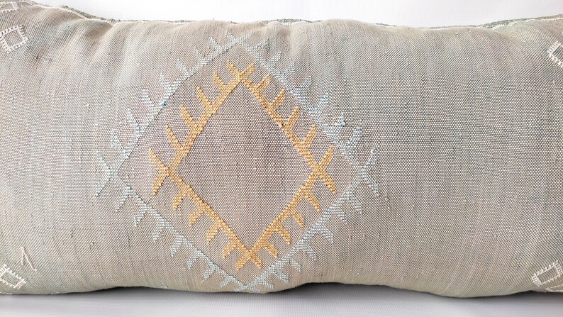 May include: Rectangular decorative pillow in a neutral gray color. The pillow features a central diamond pattern in gold and light blue, with additional geometric designs. The pillow is made of a textured fabric.