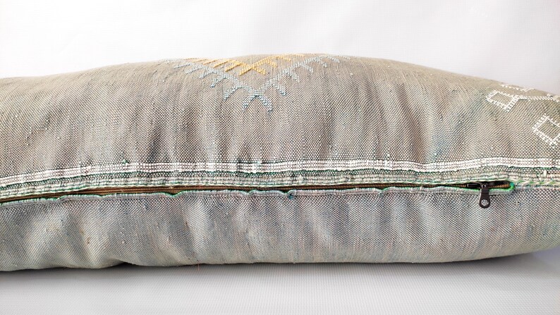 May include: A long, rectangular decorative pillow in shades of gray with embroidered geometric patterns in yellow, white, and blue. The pillow has a zipper closure and a textured fabric.