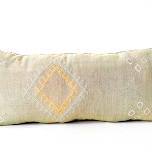 May include: A rectangular decorative pillow with a light green and beige fabric. The pillow features a central diamond pattern in yellow and white, with additional diamond shapes scattered across the surface. The edges have a green fringe.