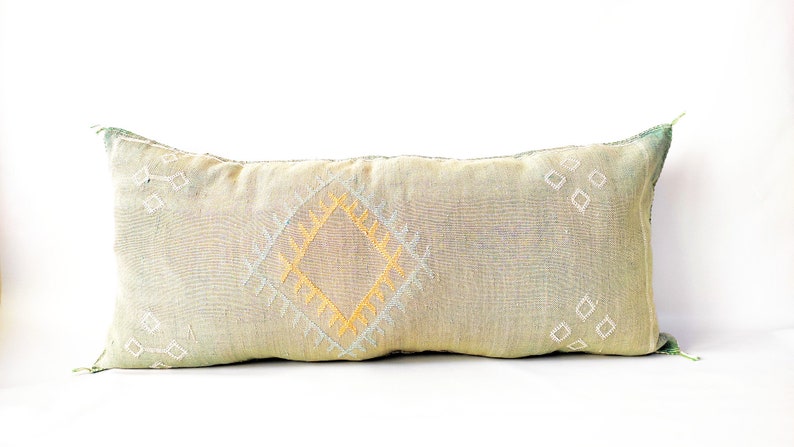 May include: A rectangular decorative pillow with a light green and beige fabric. The pillow features a central diamond pattern in yellow and white, with additional diamond shapes scattered across the surface. The edges have a green fringe.