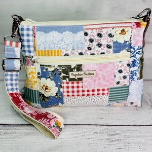May include: A rectangular fabric crossbody bag featuring a patchwork design of various floral, gingham, and lace patterns in blue, red, yellow, and white. It includes a front zippered pocket, a top zipper, and a detachable strap with the text Payneless Creations.