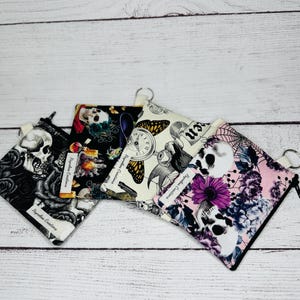 May include: Four small, square zippered pouches with various gothic-themed patterns. Designs include skulls, roses, butterflies, clocks, and floral arrangements. Each pouch has a black zipper and a small silver key ring.