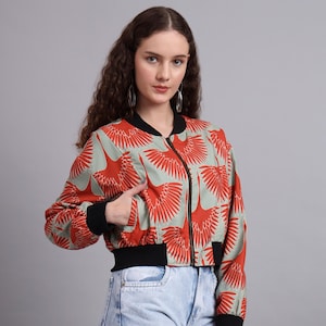 Puede incluir: Chaqueta bomber con un estampado de grullas rojas sobre un fondo verde claro. La chaqueta tiene cremallera, puños y cintura negros. Las mangas son largas y la chaqueta es corta.