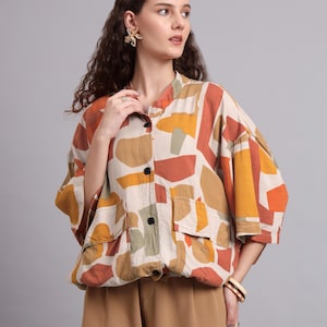 Op de afbeelding: Een shirt met korte mouwen en een abstract patroon in beige, oranje, bruin en groen. Het shirt heeft twee voorzakken en zwarte knopen. De persoon draagt een gouden oorbel en een bruine armband. Onderdeel van een kledingcollectie.