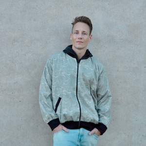 Könnte beinhalten: Hellgrüne Jacke mit Reißverschluss und Wellenmuster, schwarzem Besatz und Kapuze. Die Jacke hat einen Frontreißverschluss und eine Tasche auf der linken Seite. Die Person trägt hellblaue Jeans.