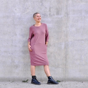 Peut inclure: Une femme portant une robe rose à manches longues avec des poches et des bottes noires. Elle se tient devant un mur en béton.