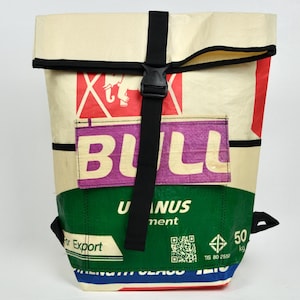 Può includere: Uno zaino con chiusura a rullo realizzato in materiale intrecciato riciclato con una combinazione di colori crema, verde e rosso. La parte anteriore mostra la scritta BULL in grandi lettere bianche su una toppa rettangolare viola e una cinghia.