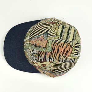 May include: A patterned baseball cap with a dark blue brim. The cap features a patchwork design with animal print and geometric patterns in shades of beige, brown, and black. The hat is set against a white background.