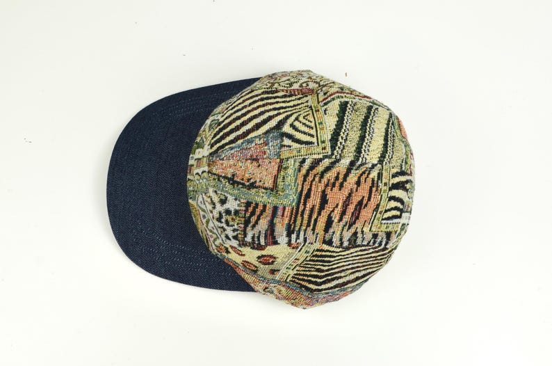 May include: A patterned baseball cap with a dark blue brim. The cap features a patchwork design with animal print and geometric patterns in shades of beige, brown, and black. The hat is set against a white background.
