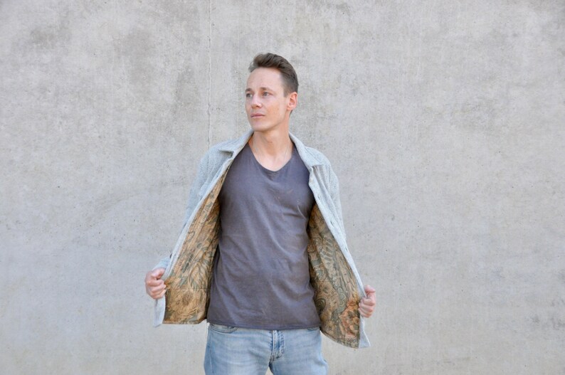 Puede incluir: Un hombre con una chaqueta gris con un forro estampado. La chaqueta est&aacute; desabrochada y el forro es visible. Tambi&eacute;n lleva una camiseta gris y unos vaqueros azules.