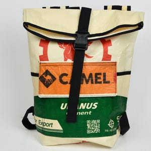 Könnte beinhalten: Ein Rolltop-Rucksack aus cremefarbenem, orangefarbenem und grünem Recyclingmaterial. Die Vorderseite verfügt über einen schwarzen vertikalen Riemen mit Kunststoffschnalle, einen orangefarbenen rechteckigen Aufnäher mit dem Text CAMEL und grüne Abschnitte mit dem Text U NUS.