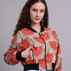 Könnte beinhalten: Eine Bomberjacke mit rotem Kranichmuster auf hellgrünem Hintergrund. Die Jacke hat einen schwarzen Kragen, Bündchen und Saum sowie einen schwarzen Reißverschluss. Die Jacke ist im Cropped-Stil gehalten.