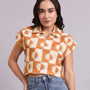 Könnte beinhalten: Kurzärmliges Crop-Top mit Kragen in einem Schachbrettmuster aus Orange und Creme. Die Quadrate weisen ein Sonnenstrahl-Design auf. Das Oberteil wird mit einer blauen Jeans kombiniert.