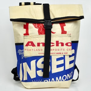 Könnte beinhalten: Ein Rolltop-Rucksack aus wiederverwertetem Material in Creme, Blau und Rot. Die Vorderseite zeigt eine Tasche mit dem Text Ancho Portland Composite Cement und einen großen blauen Bereich mit dem Text Insee Diamond. Ein schwarzer Riemen mit Schnalle sichert das Oberteil.