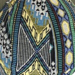 May include: Close-up of a baseball cap with a detailed geometric pattern. The cap features a mix of colors, including yellow, blue, green, and black, creating a vibrant and textured design. The cap's brim is a solid olive green.