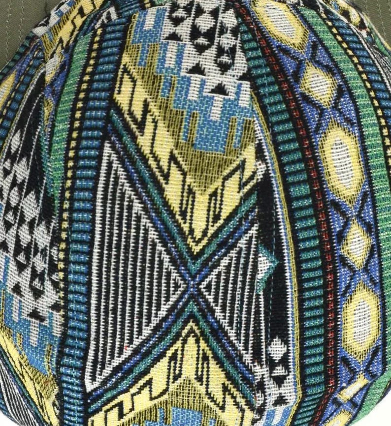 May include: Close-up of a baseball cap with a detailed geometric pattern. The cap features a mix of colors, including yellow, blue, green, and black, creating a vibrant and textured design. The cap's brim is a solid olive green.