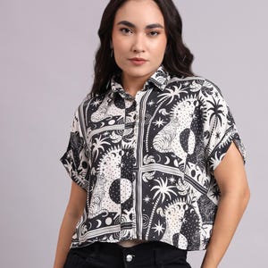 Puede incluir: Camisa abotonada con estampado en blanco y negro con mangas cortas. La camisa presenta un diseño repetido de tigres, lunas, estrellas y palmeras. La camisa es corta y tiene cuello.