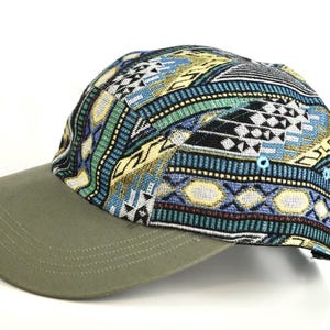 May include: A five-panel cap with a patterned top and a solid olive green brim. The top features a colorful geometric design in blue, green, yellow, and black. The cap has a curved brim and a structured fit.