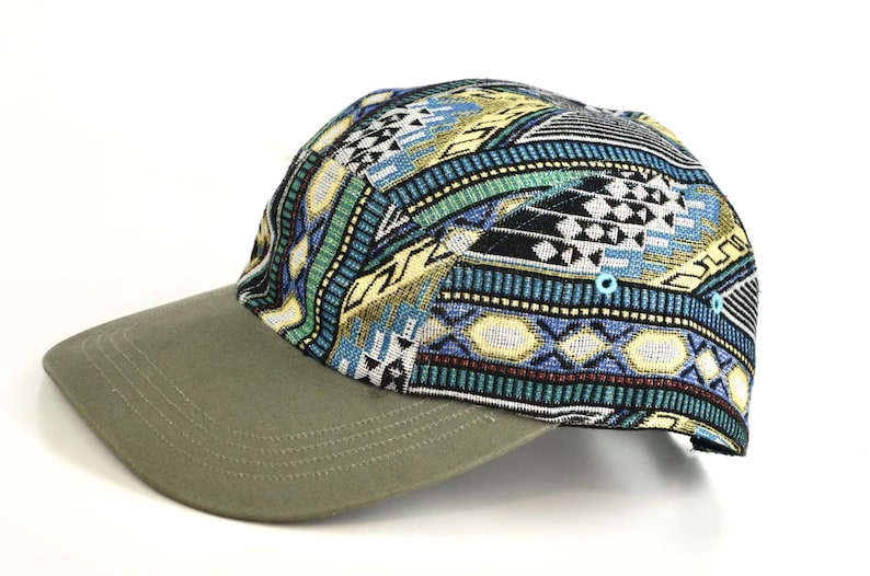 May include: A five-panel cap with a patterned top and a solid olive green brim. The top features a colorful geometric design in blue, green, yellow, and black. The cap has a curved brim and a structured fit.