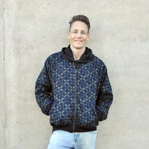 Könnte beinhalten: Ein Mann trägt eine Jacke mit blauem und grünem Muster und Reißverschluss. Die Jacke hat eine Kapuze und der Mann steht vor einer grauen Wand.