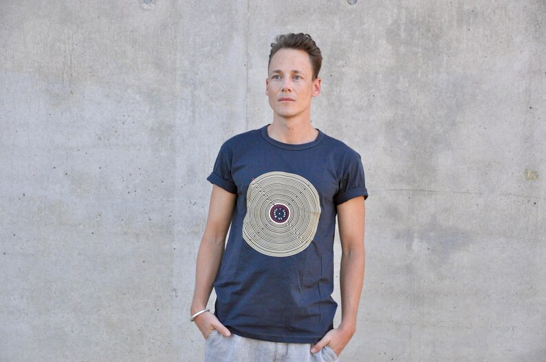 Pu&ograve; includere: Una t-shirt blu scuro con un design circolare in beige e marrone. Il design presenta cerchi concentrici e un cerchio pi&ugrave; piccolo al centro con un motivo a stella.