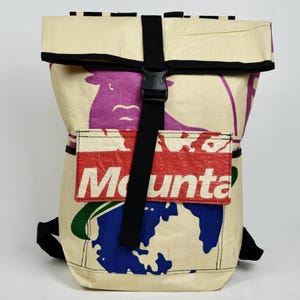 Könnte beinhalten: Ein Rolltop-Rucksack aus cremefarbenem Material mit einem Grafikdruck, der eine violette Tiersilhouette, eine blaue Weltkugelform und eine rote rechteckige Tasche mit dem weißen Text "Mounta" auf der Vorderseite zeigt, gesichert durch einen schwarzen Riemen.