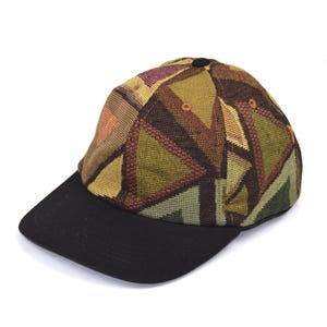 May include: A baseball cap featuring a crown with a woven geometric triangle pattern in shades of olive green, brown, and muted orange. The cap has a solid black brim and a small black button at the top center of the crown, with visible eyelets.