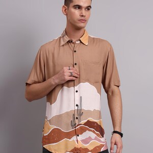 Könnte beinhalten: Kurzärmeliges Hemd in Beige mit Wüstenlandschaftsdesign. Das Hemd hat eine Brusttasche und einen Kragen mit Kontrastfarbe. Die Wüstenszene zeigt einen Kaktus und Erdtöne.