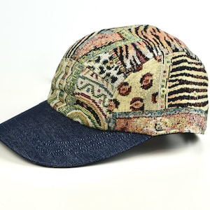 May include: A baseball cap with a denim blue brim and a patterned crown. The crown features a patchwork design with various patterns, including animal prints and geometric shapes, in shades of beige, brown, and green. The cap is set against a white background.