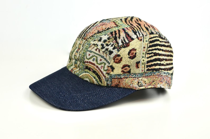 May include: A baseball cap with a denim blue brim and a patterned crown. The crown features a patchwork design with various patterns, including animal prints and geometric shapes, in shades of beige, brown, and green. The cap is set against a white background.