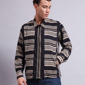 May include: A black and beige striped zip-up shirt. The shirt features horizontal stripes and a vertical design down the centre. The zip is black, and the shirt has long sleeves and a collar. The shirt is worn with blue jeans.
