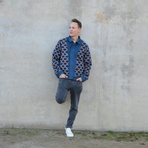 Peut inclure: Un homme portant une veste en jean bleu avec un motif g&eacute;om&eacute;trique sur le devant et le dos. Il porte un jean gris fonc&eacute; et des baskets blanches. Il se tient devant un mur en b&eacute;ton.