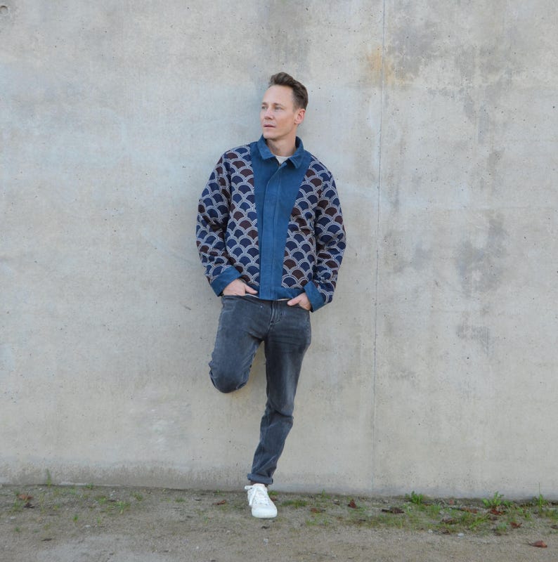 Peut inclure: Un homme portant une veste en jean bleu avec un motif g&eacute;om&eacute;trique sur le devant et le dos. Il porte un jean gris fonc&eacute; et des baskets blanches. Il se tient devant un mur en b&eacute;ton.