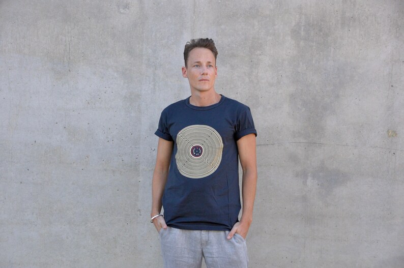Pu&ograve; includere: Una t-shirt blu navy con un design circolare in oro e marrone. Il design presenta cerchi concentrici con un cerchio pi&ugrave; piccolo al centro.